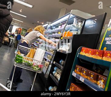 Vue arrière du chariot de magasinage jeune femme permettant d'accéder à l'étagère de réfrigération de yogourt dans l'allée du magasin M&S Marks & Spencer 2024 R.-U. KATHY DEWITT Banque D'Images