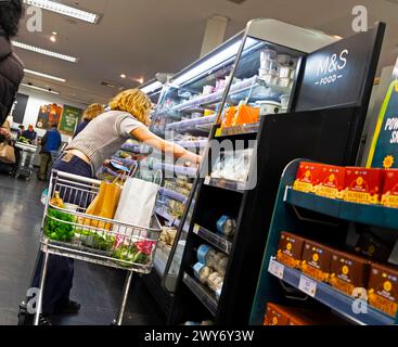 Chariot de magasinage jeune femme vue arrière atteignant le yogourt sur l'étagère de réfrigération M&S Marks & Spencer allée intérieure 2024 UK KATHY DEWITT Banque D'Images