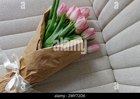 Bouquet de tulipes roses pastel fraîches enveloppées dans le papier artisanal décoré d'un ruban de satin couché sur la chaise beige, Banque D'Images