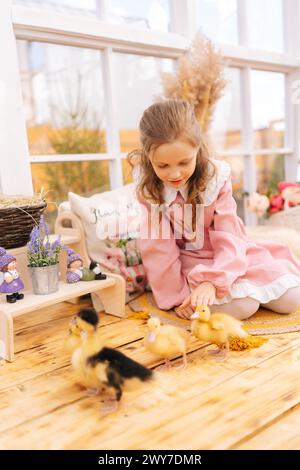 Portrait vertical de mignonne petite fille en robe jouant avec de petits canetons jaunes se trouvant sur le sol de la maison gazebo d'été le jour ensoleillé. Banque D'Images