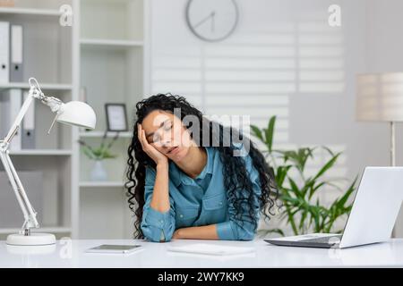 Une femme d'affaires surchargée de travail faisant une sieste dans son espace de travail, avec un ordinateur portable et un téléphone, dans un cadre de bureau moderne. Banque D'Images