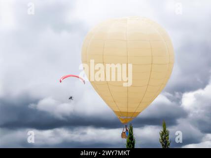 Grande montgolfière volant dans le ciel avec des nuages à côté d'un paramoteur lors d'un festival aérien. Banque D'Images