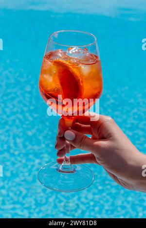 Aperol Spritz cocktail alcoolisé dans un gobelet en verre dans la main de la femme avec vernis à ongles blanc sur fond de piscine d'eau bleue Banque D'Images