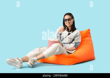 Belle jeune femme heureuse avec seau de pop-corn regardant la télévision sur la chaise Beanbag sur fond bleu Banque D'Images