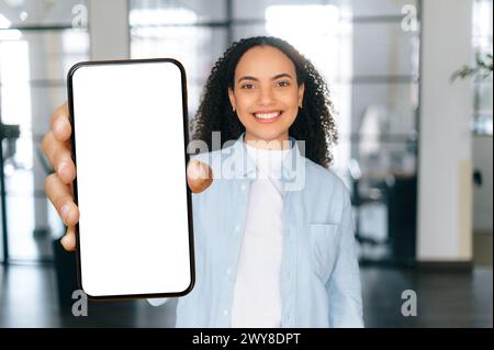 Jeune femme aux cheveux bouclés multiraciaux étonné, se tient dans un espace de bureau moderne, montrant un téléphone intelligent avec un écran blanc vide de maquette, regarde la caméra, sourire heureux. Espace de copie, concept de maquette Banque D'Images