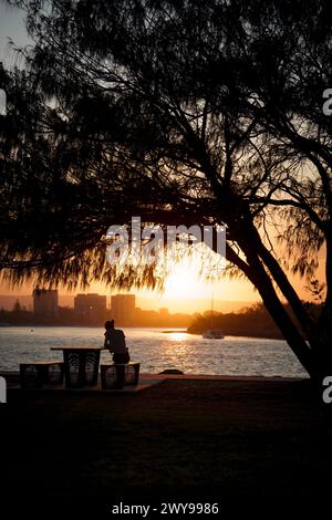 Une figure solitaire se penche soigneusement sur un banc au bord de la rivière au coucher du soleil, encadrée par des arbres silhouettes et une lumière dorée se reflétant sur l'eau tranquille. Banque D'Images