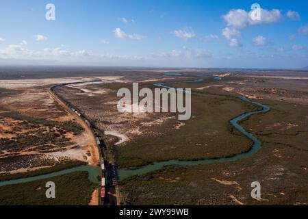 Vue aérienne d'un train de marchandises intermodal passant au-dessus d'une rivière sinueuse à travers un paysage sec avec une végétation clairsemée sous un ciel bleu Banque D'Images