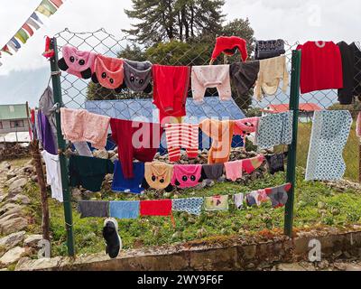 Vêtements colorés accrochés à une clôture métallique avec des drapeaux de prière et des arbres en arrière-plan. Banque D'Images