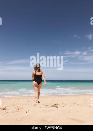 Femme en maillot de bain noir marchant vers la mer Banque D'Images