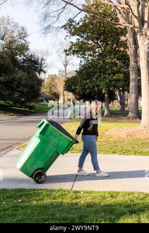 Femme roulant un bac de recyclage vert sur un trottoir. Banque D'Images