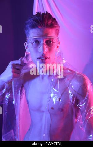 Jeune homme élégant portant un manteau transparent et des lunettes dans les néons Banque D'Images