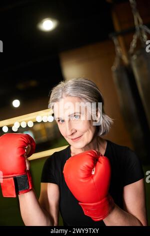 belle femme senior joyeuse dans les vêtements de sport avec les cheveux gris posant avec des gants de boxe pendant que dans la salle de gym Banque D'Images