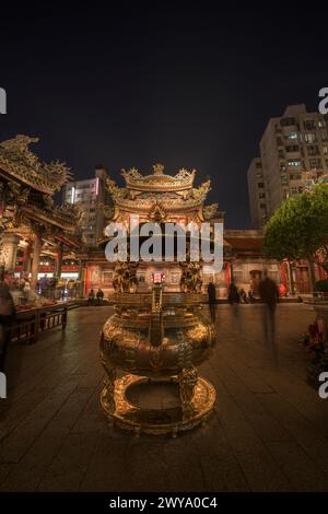 La photo montre une porte d'entrée du temple Longshan richement décorée avec des sculptures complexes et animée de fidèles la nuit Banque D'Images