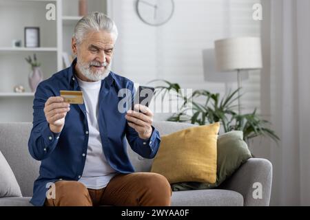 Homme barbu mûr confortablement utilisant son smartphone et sa carte de crédit pour magasiner en ligne de la maison. Banque D'Images