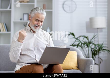 Vieux gentleman utilisant un ordinateur portable et montrant l'excitation avec un geste gagnant dans un cadre de salon confortable. Banque D'Images