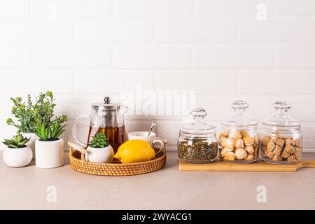 Fond de cuisine élégant avec théière en verre, citron, tasse, pots de thé, sucre, pâtisserie meringue. Plantes vertes en pots. Vue de face Banque D'Images