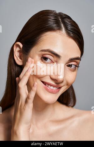 Une jeune femme caucasienne aux cheveux bruns souriant alors qu'elle touche doucement sa joue avec de la crème. Banque D'Images