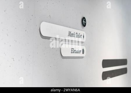 Mur en béton gris avec panneaux directionnels indiquant les toilettes pour hommes et l'ascenseur Banque D'Images