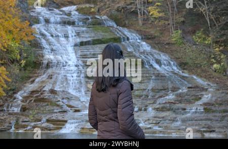 femme regardant une cascade sur une randonnée dans la nature (photographiée de derrière) portant chapeau d'hiver, veste brune (buttermilk falls parc d'état à ithaca new y Banque D'Images