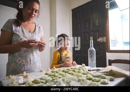 Authentique belle jeune femme, mère et son petit enfant, fille dans la cuisine rurale, sculptant des boulettes de pâte avec remplissage de purée de pommes de terre Banque D'Images