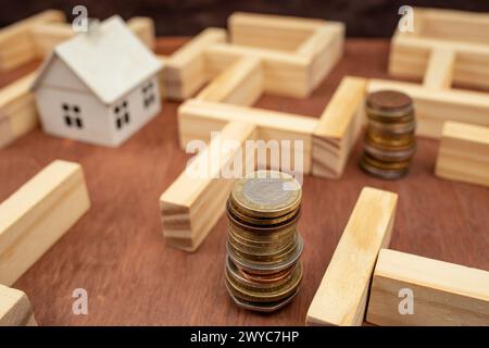 Piles de pièces de monnaie et une maison blanche miniature dans un labyrinthe en bois, métaphore des difficultés économiques Banque D'Images