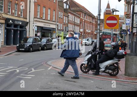 Homme en veste bleue traversant la route près d'une station de taxis alors qu'un conducteur est assis sur un scooter, Reading Banque D'Images