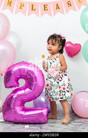 Petite fille brune latine tenant un bonbon dans ses mains, dans le fond des ballons d'anniversaire et un grand numéro deux Banque D'Images