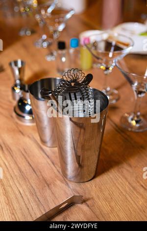 Une vue intime du shaker en acier inoxydable d'un barman et des outils de mélange sur un bar en bois, avec des verres à cocktail en arrière-plan. Banque D'Images