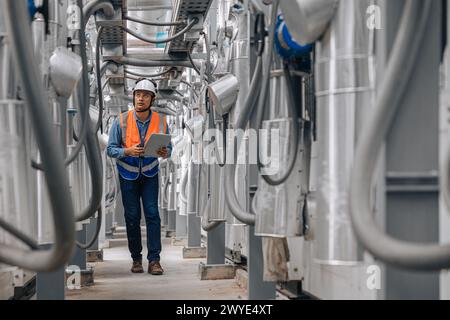 l'homme dans un gilet de sécurité marche à travers un grand bâtiment industriel avec des tuyaux et des fils. Il tient une planche à pince inspectant la zone du pipeline. Banque D'Images