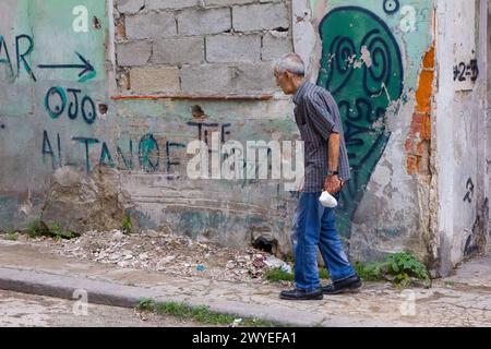 Cubain senior avec un petit sac marche sur un trottoir cassé et un mur avec des graffitis à la Havane, Cuba Banque D'Images