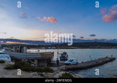 Mellieha, Malte - 21 décembre 2022 : un bateau de pêche sur terre près d'une petite jetée à Mellieha Bay pendant le coucher du soleil, en arrière-plan est Marfa Ridge. Banque D'Images