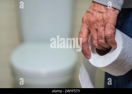 La main d'un homme plus âgé tenant un rouleau de papier toilette devant les toilettes. Banque D'Images