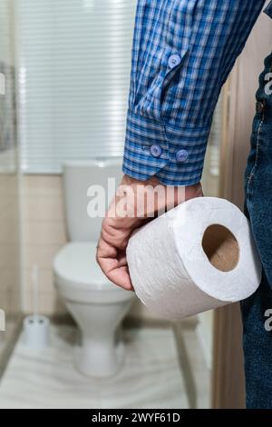 La main d'un homme plus âgé tenant un rouleau de papier toilette devant les toilettes. Banque D'Images