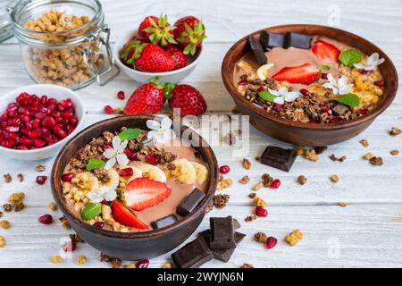 Bol à smoothie au chocolat surmonté de granola d'avoine, banane, fraises, grenade et fleurs de printemps sur une table en bois blanc. Protéines végétaliennes saines Banque D'Images