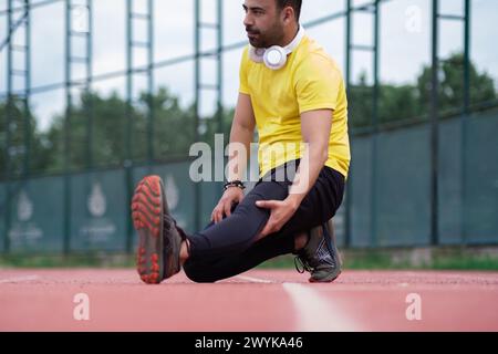 Athlète en vêtements d'activité et écouteurs étire la jambe, sur la piste de course , échauffement avant l'entraînement Banque D'Images