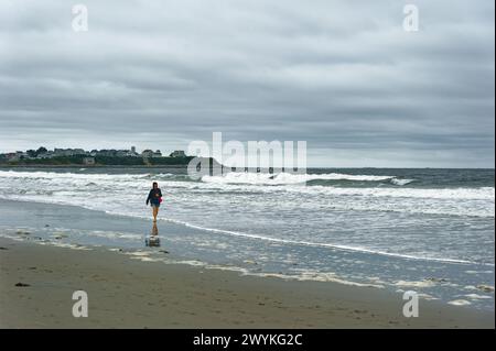 2023 Hampton Beach, New Hampshire. Une femme portant un short et un manteau d'automne marche le long de la plage un jour d'automne couvert. Banque D'Images