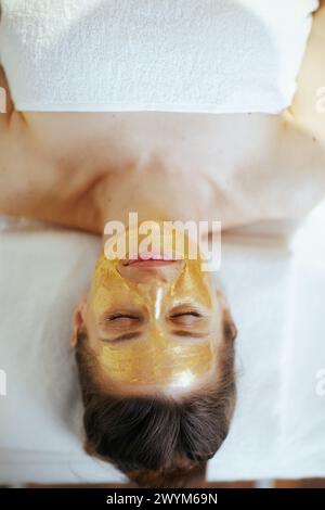 Le temps des soins de santé. Vue supérieure de la femme moderne détendue dans l'armoire de massage avec masque cosmétique doré sur le visage posé sur la table de massage. Banque D'Images