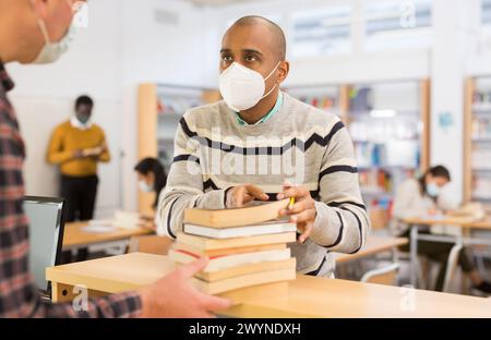 Étudiant hispanique adulte dans le masque protecteur retournant des livres au bibliothécaire Banque D'Images