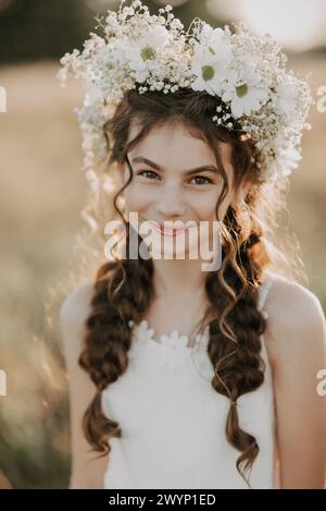 portrait d'une jeune fille souriante dans une robe blanche et une couronne florale sur ses cheveux avec des tresses dans le champ d'été. Style boho. Ajouté un petit grain, im Banque D'Images