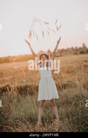 Fille heureuse en robe blanche joue et profite de l'été dans le champ au coucher du soleil. Ajout d'une simulation de petit grain de la photographie de film Banque D'Images