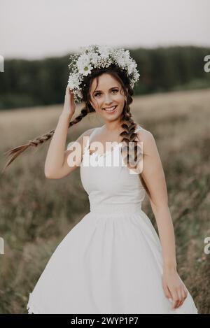 joyeuse fille heureuse avec des tresses et une couronne de fleurs dans une robe blanche sourit joyeusement et a de la joie dans le champ d'été. Style de vie bohémien. Ajout d'un petit Banque D'Images