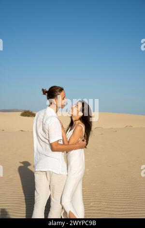 Vue verticale de jeune couple en tenue blanche se regardant dans les dunes de sable à la plage avec ciel dégagé en été Banque D'Images