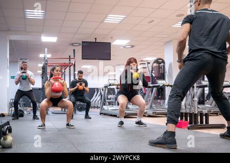 Un groupe diversifié de sportifs s'engage dans une séance de squat kettlebell avec un coaching personnalisé d'un instructeur de fitness dans un gymnase bien équipé. Banque D'Images