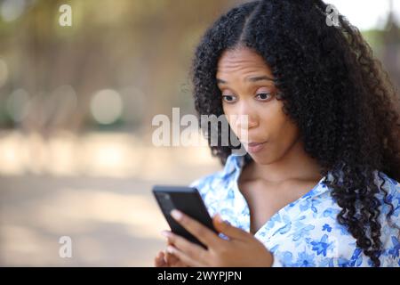 Femme noire étonnée vérifiant le contenu du téléphone debout à l'extérieur Banque D'Images