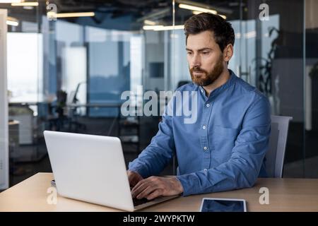 Homme professionnel sérieux dans une chemise bleue profondément concentré sur le travail à l'aide d'un ordinateur portable dans un cadre de bureau d'entreprise lumineux. Banque D'Images