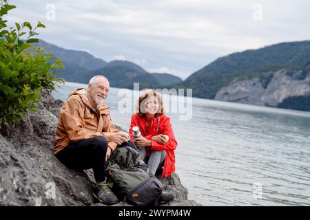 Couple de personnes âgées actives randonnant ensemble dans les montagnes. Boire du café et avoir une collation saine, pour obtenir de l'énergie. Touriste senior avec des sacs à dos au repos Banque D'Images