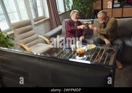 Santé. Hommes âgés, amis assis sur le canapé à la maison, boire de la bière, manger des collations et regarder la traduction de match de sport en ligne Banque D'Images