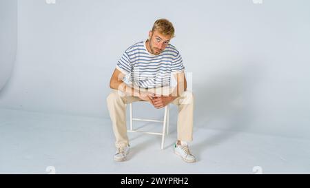 bel homme blond regardant droit avec des vêtements décontractés sur fond blanc, assis sur une chaise avec les mains pliées. Concept de mode Banque D'Images