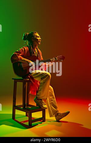 Jeune homme talentueux, musicien avec dreadlock assis sur la chaise et jouant de la guitare sur fond vert rouge dégradé dans la lumière néon Banque D'Images