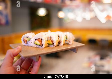 Une main tient une assiette de rouleaux de sushi au saumon sur un décor de restaurant japonais chaud et coloré. Le bokeh et le flou d'arrière-plan sont très beaux Banque D'Images